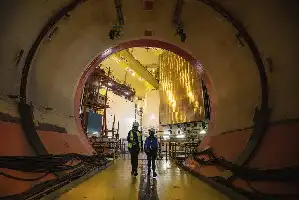Description de l'image par IA : Deux ouvriers en combinaison de sécurité marchent dans un tunnel industriel bien éclairé avec des câbles au sol.