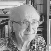 Description de l'image par IA : Photo en noir et blanc d'une personne âgée portant des lunettes, souriant, devant une étagère de livres.